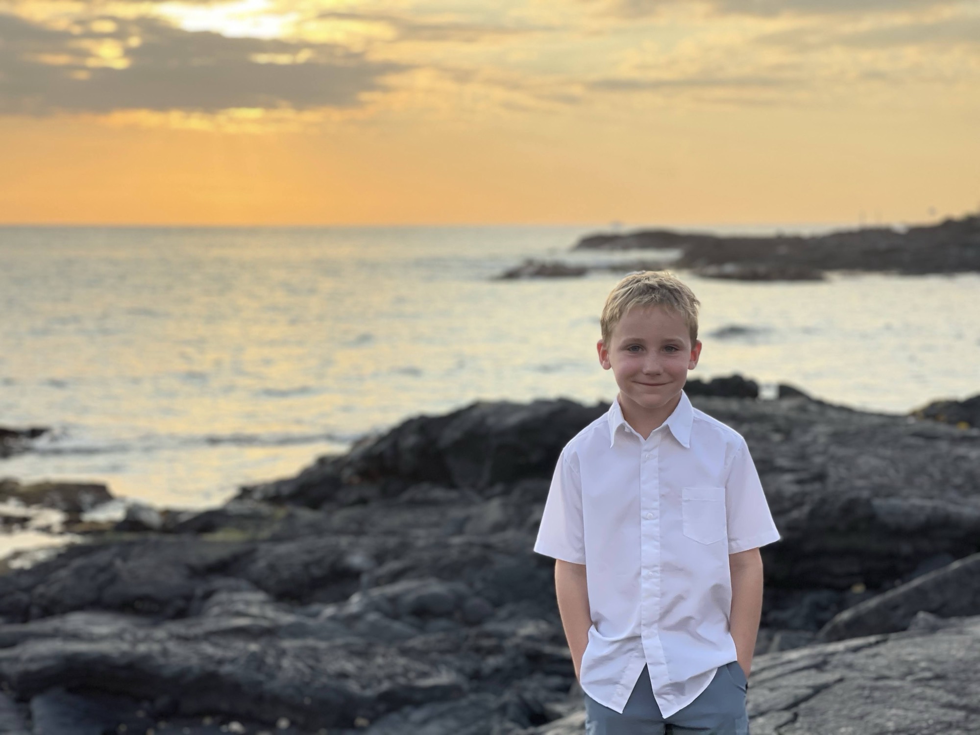 A young boy in a white short-sleeved button-down shirt stands smiling with his hands in his pockets on a rocky shoreline. The ocean and a golden sunset sky are visible in the background.