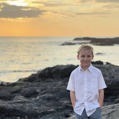 A young boy in a white short-sleeved button-down shirt stands smiling with his hands in his pockets on a rocky shoreline. The ocean and a golden sunset sky are visible in the background.