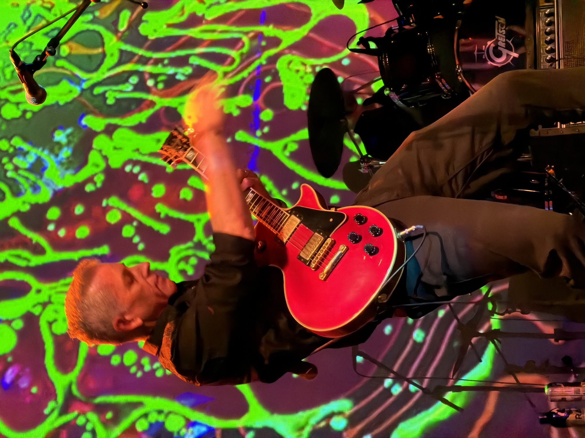 A guitarist plays a red electric guitar on stage during a live performance, with a drummer visible in the background. The stage is illuminated by a vibrant, psychedelic green and red light projection.