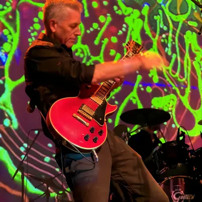 A guitarist plays a red electric guitar on stage during a live performance, with a drummer visible in the background. The stage is illuminated by a vibrant, psychedelic green and red light projection.