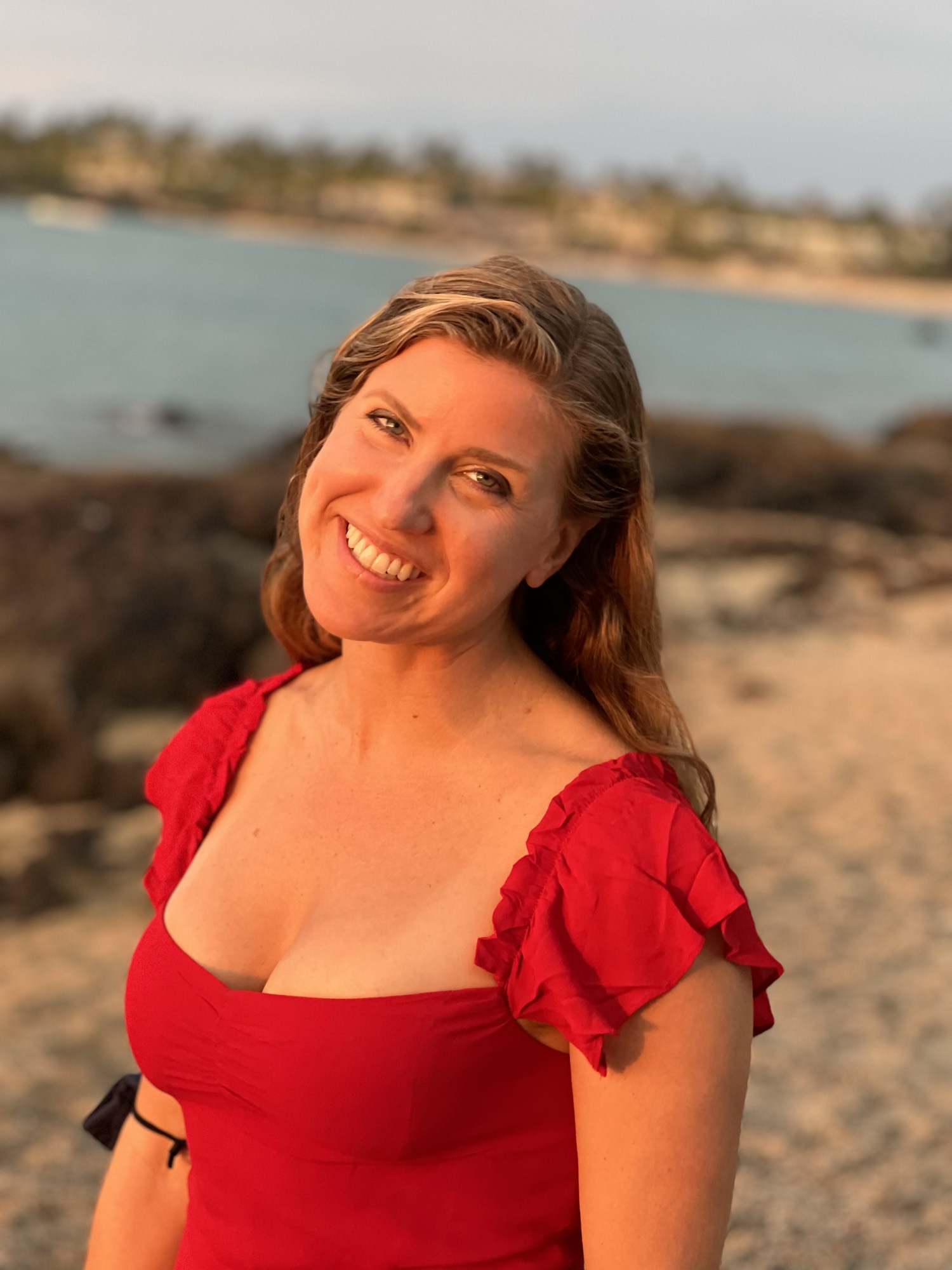 A smiling woman in a red top poses for a portrait on a sandy beach during sunset, with the ocean and a rocky shoreline visible in the background.