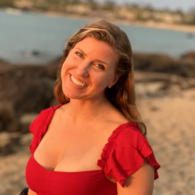 A smiling woman in a red top poses for a portrait on a sandy beach during sunset, with the ocean and a rocky shoreline visible in the background.