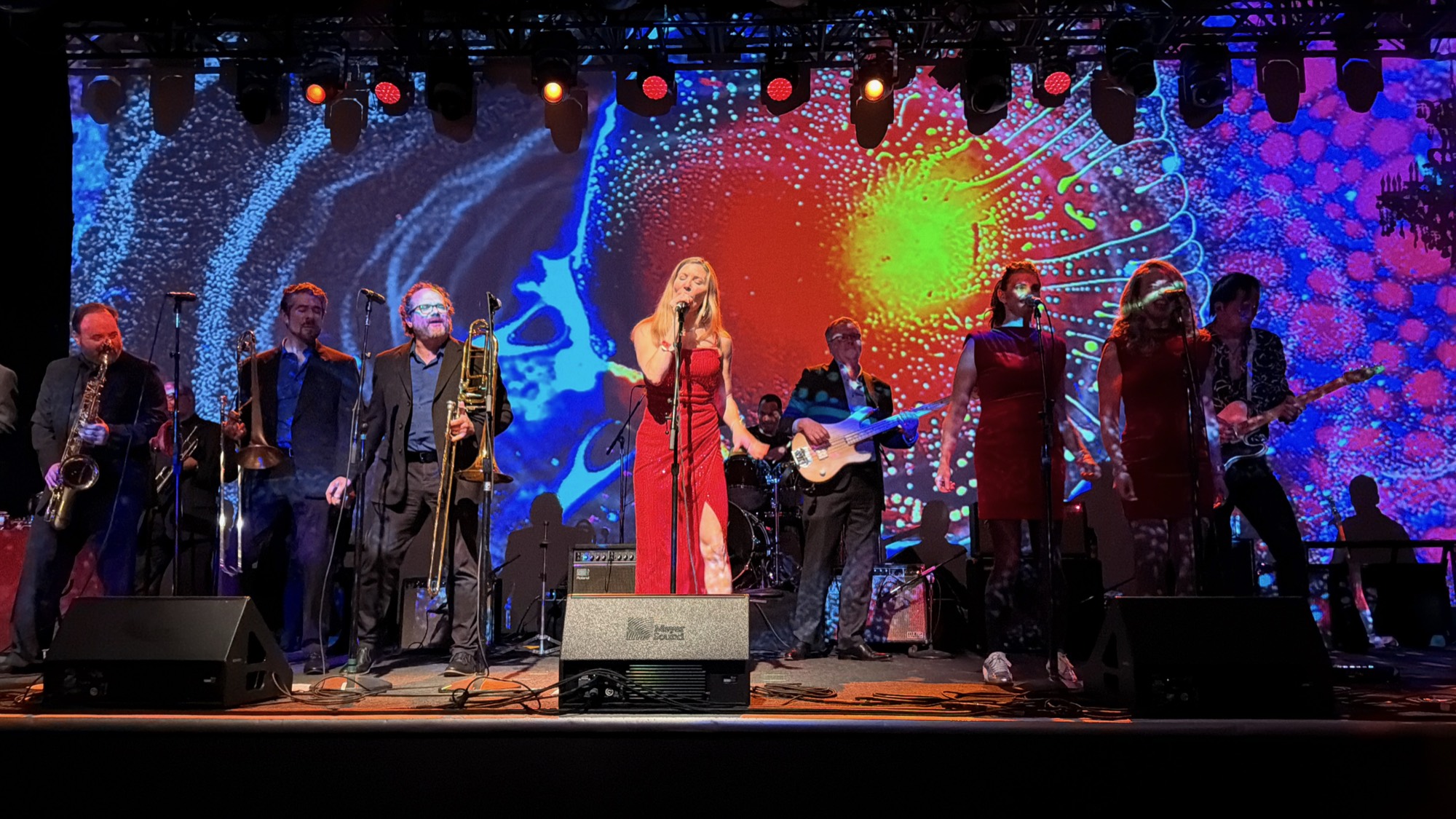 A musical band performs live on a stage featuring a large, colorful, abstract projection screen in the background. The group includes a female lead singer in a red dress, backup singers, a brass section, a bassist, and a guitarist.
