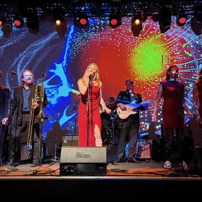 A musical band performs live on a stage featuring a large, colorful, abstract projection screen in the background. The group includes a female lead singer in a red dress, backup singers, a brass section, a bassist, and a guitarist.