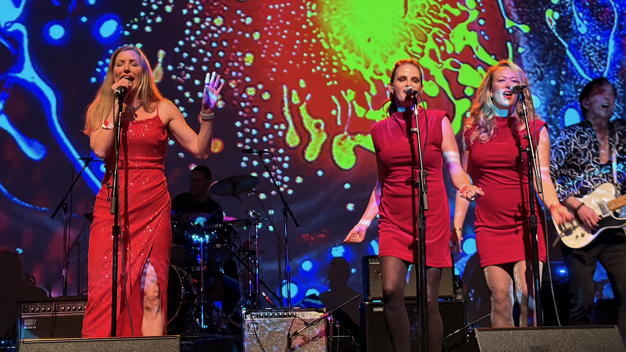 Three female vocalists in red dresses sing into microphones on a stage, backed by a drummer and a guitarist. They are performing in front of a large screen displaying a vibrant, multicolored psychedelic pattern.