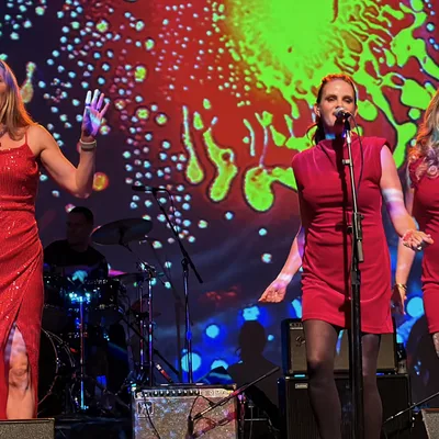 Three female vocalists in red dresses sing into microphones on a stage, backed by a drummer and a guitarist. They are performing in front of a large screen displaying a vibrant, multicolored psychedelic pattern.
