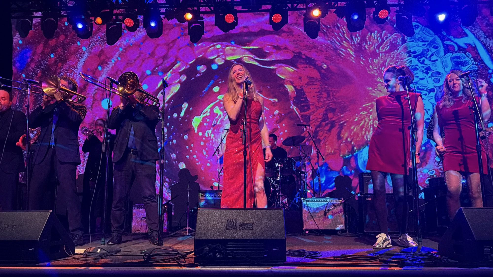 A musical band performs live on a stage featuring a vibrant, colorful backdrop and stage lighting. The group includes a female lead singer in a red dress, two female backup singers, a brass section with a trombone and trumpet, and a drummer in the background.