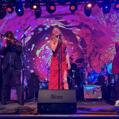 A musical band performs live on a stage featuring a vibrant, colorful backdrop and stage lighting. The group includes a female lead singer in a red dress, two female backup singers, a brass section with a trombone and trumpet, and a drummer in the background.