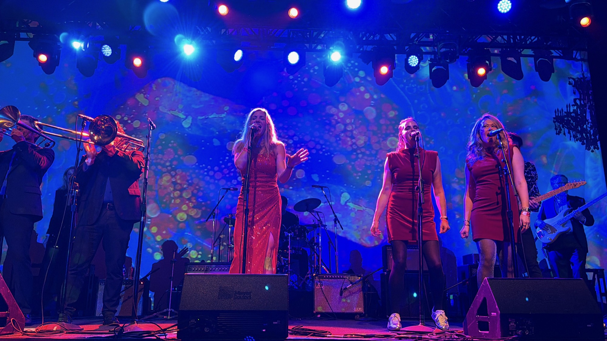 A musical band performs on a stage illuminated by vibrant blue, red, and purple lights, with three female vocalists in red dresses singing at microphones in the foreground. They are accompanied by a brass section on the left and a guitarist and bassist on the right, set against a colorful, abstract backdrop.