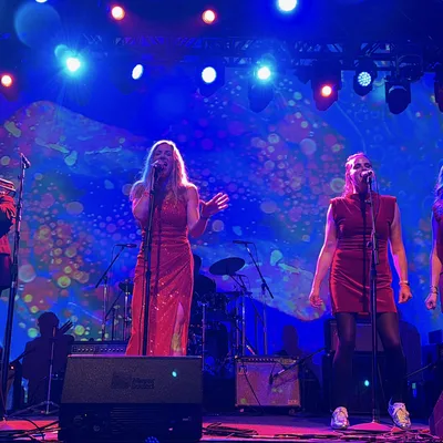 A musical band performs on a stage illuminated by vibrant blue, red, and purple lights, with three female vocalists in red dresses singing at microphones in the foreground. They are accompanied by a brass section on the left and a guitarist and bassist on the right, set against a colorful, abstract backdrop.