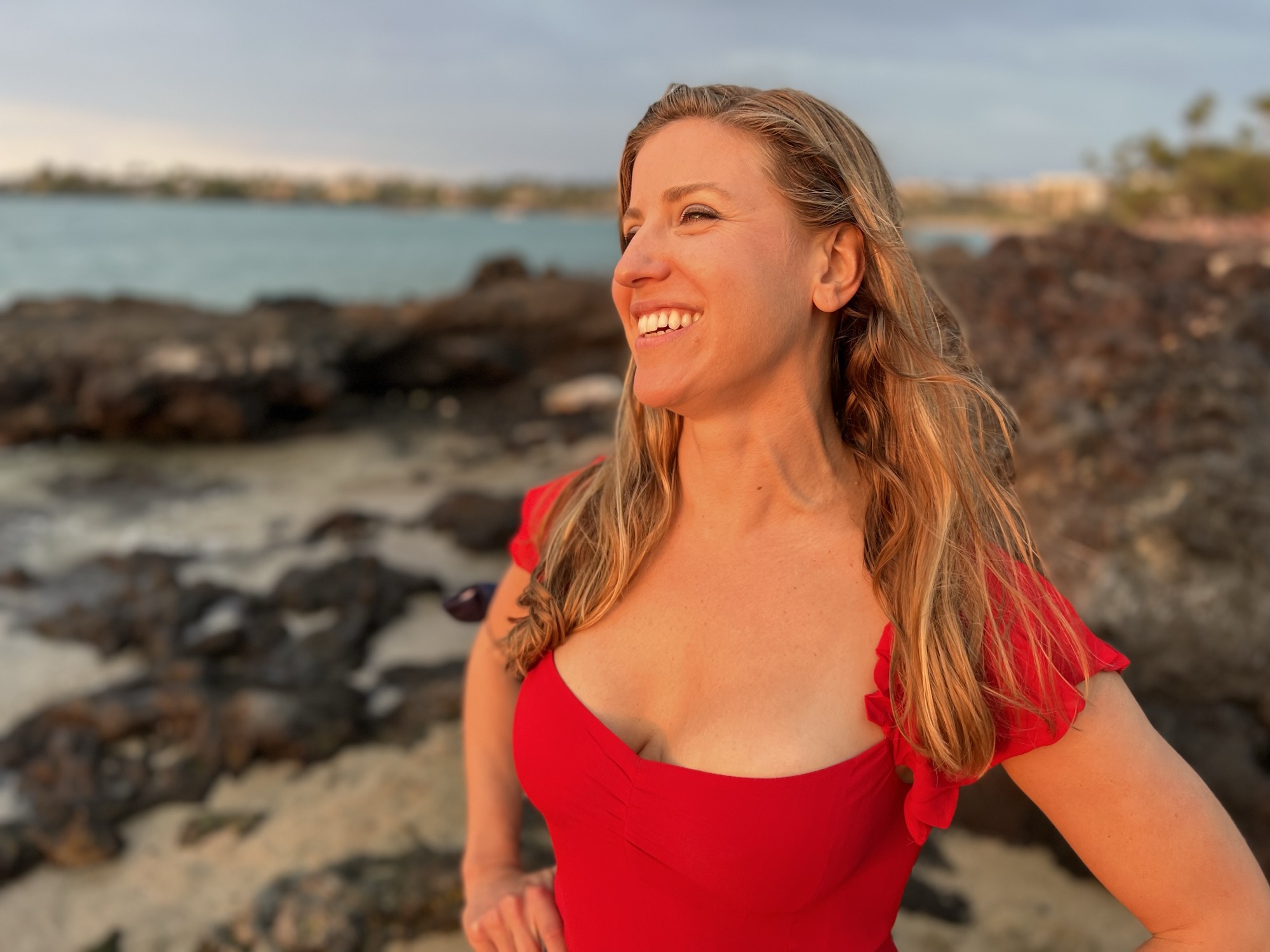 A smiling woman in a red top stands on a rocky beach, looking off-camera during sunset. The background features a body of water and a distant shoreline under a soft evening sky.
