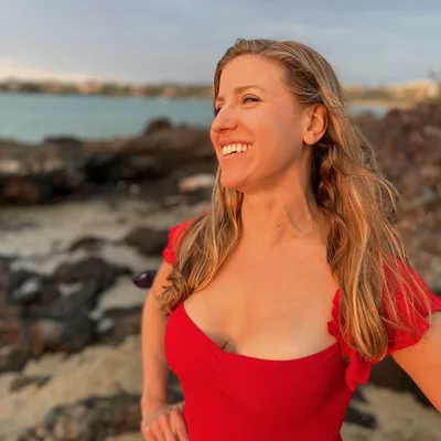 A smiling woman in a red top stands on a rocky beach, looking off-camera during sunset. The background features a body of water and a distant shoreline under a soft evening sky.