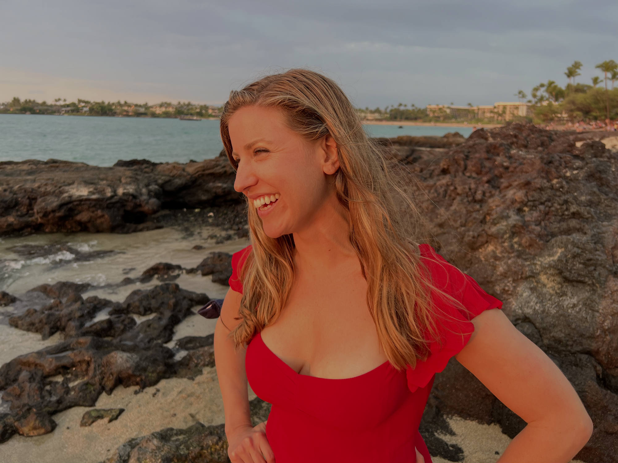 A smiling woman in a red dress stands on a rocky beach during sunset, with her hands on her hips as she looks off-camera. The background features a calm ocean, volcanic rocks, and a distant shoreline with palm trees under a cloudy sky.