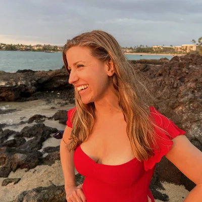A smiling woman in a red dress stands on a rocky beach during sunset, with her hands on her hips as she looks off-camera. The background features a calm ocean, volcanic rocks, and a distant shoreline with palm trees under a cloudy sky.