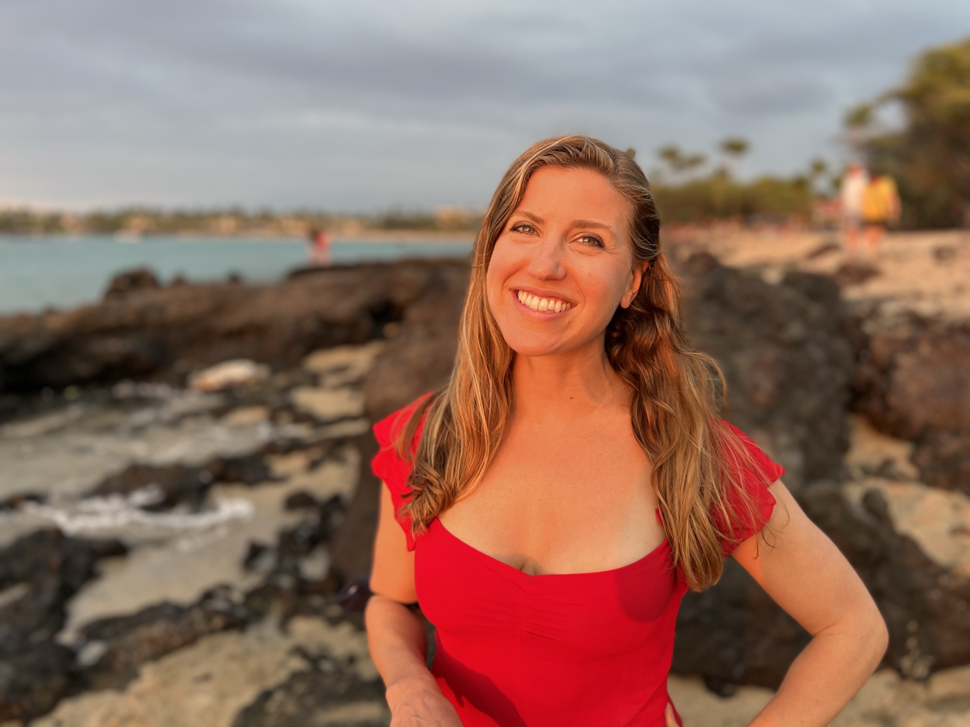 A smiling woman in a red top poses for a portrait on a rocky beach during sunset. The background features the ocean, a sandy shoreline, and a cloudy sky illuminated by the warm evening light.
