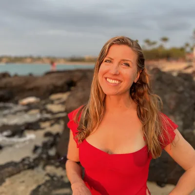 A smiling woman in a red top poses for a portrait on a rocky beach during sunset. The background features the ocean, a sandy shoreline, and a cloudy sky illuminated by the warm evening light.