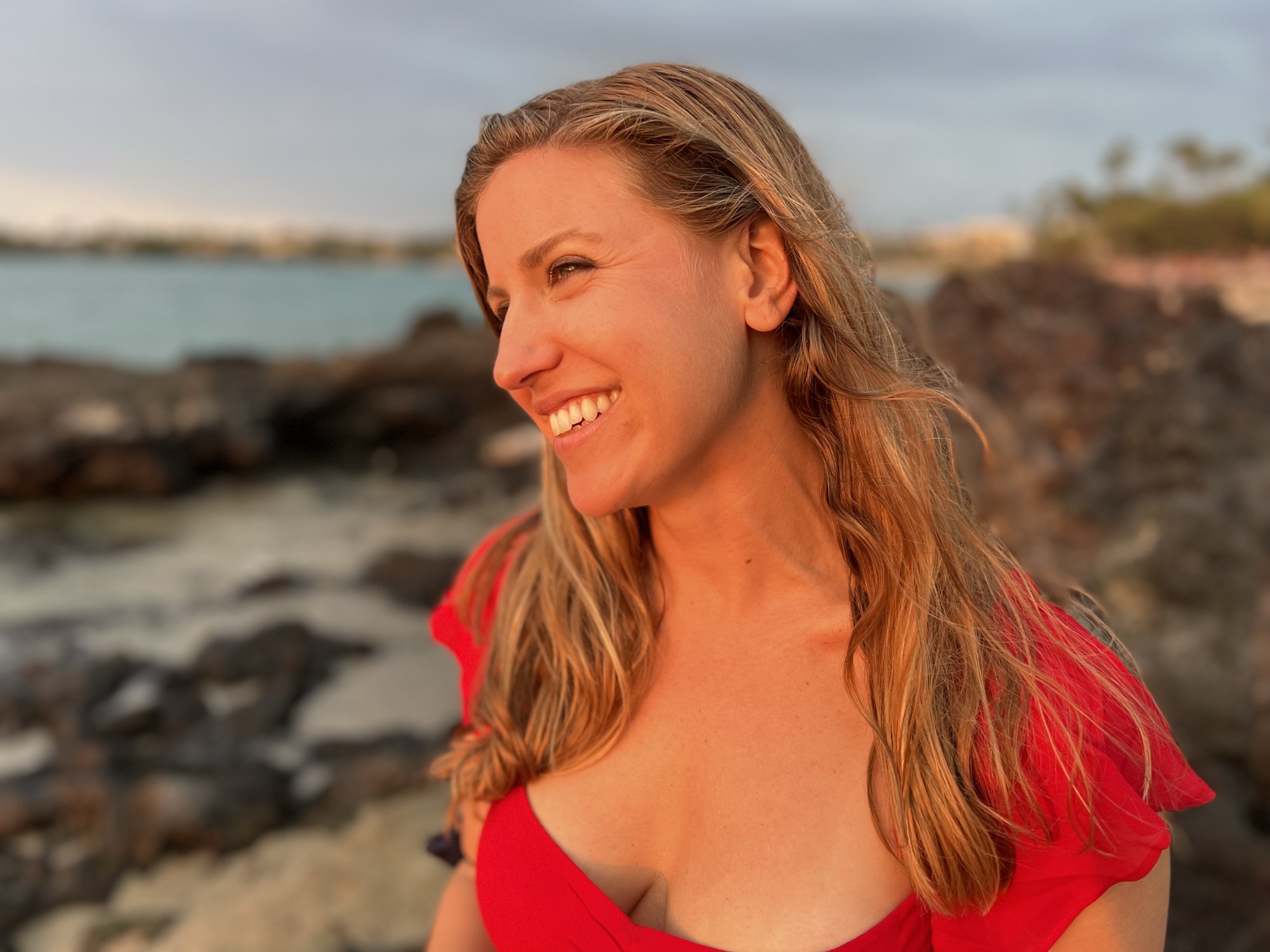 A close-up portrait shows a smiling woman in a red top looking off-camera, illuminated by the warm light of a sunset. She is standing outdoors on a rocky shoreline with the ocean visible in the background.
