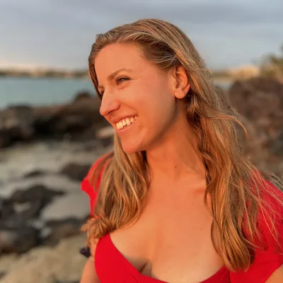 A close-up portrait shows a smiling woman in a red top looking off-camera, illuminated by the warm light of a sunset. She is standing outdoors on a rocky shoreline with the ocean visible in the background.