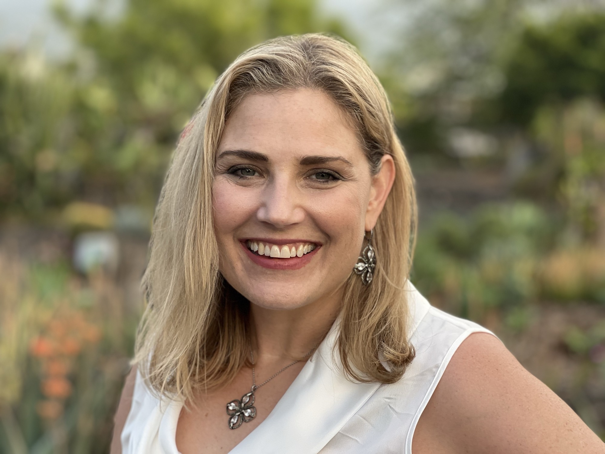 A close-up portrait shows a smiling blonde woman wearing a white sleeveless top, a matching necklace, and earrings. She is posing outdoors with a softly blurred background of green foliage and garden plants.