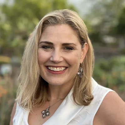 A close-up portrait shows a smiling blonde woman wearing a white sleeveless top, a matching necklace, and earrings. She is posing outdoors with a softly blurred background of green foliage and garden plants.