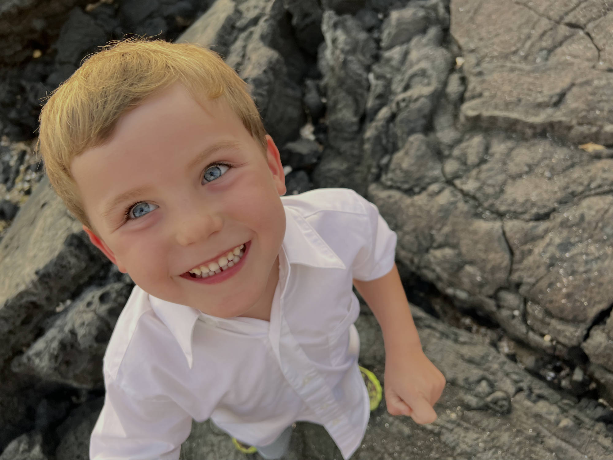 A young boy with blue eyes and a white button-down shirt smiles up at the camera. He is standing outdoors on dark, textured volcanic rocks.