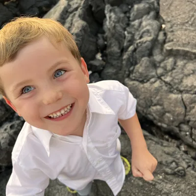 A young boy with blue eyes and a white button-down shirt smiles up at the camera. He is standing outdoors on dark, textured volcanic rocks.