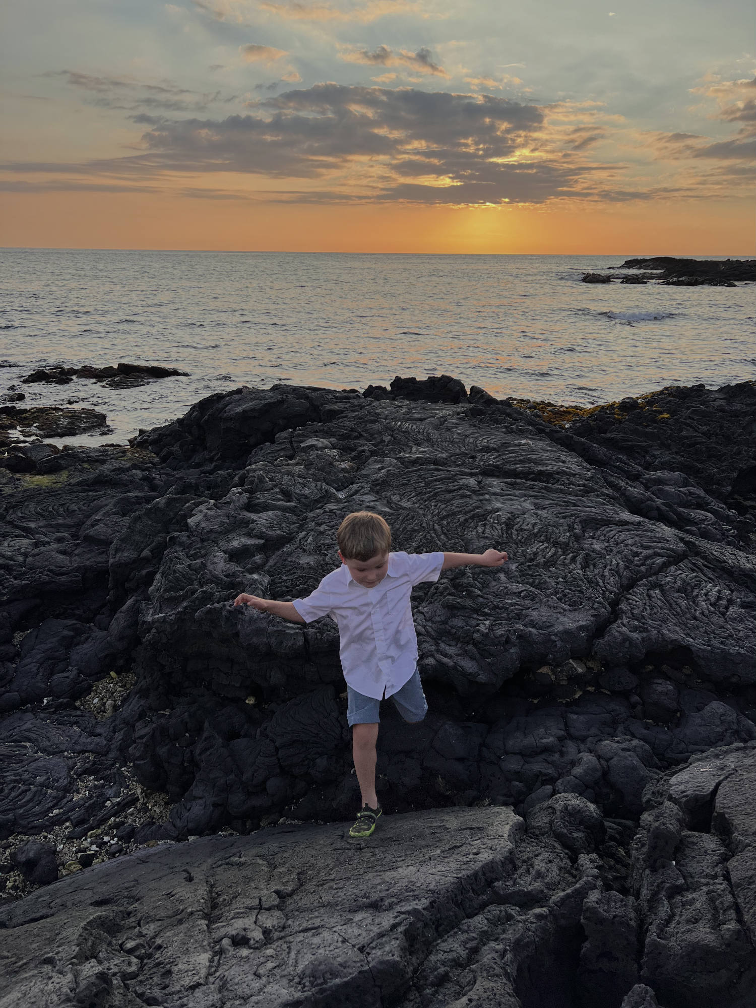 A young boy in a white shirt and blue shorts balances on one foot while navigating dark volcanic rock formations along a coastline. In the background, the sun sets over the ocean, casting a warm orange glow across the sky and water.