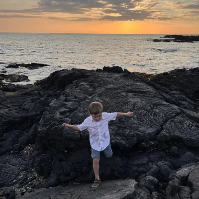 A young boy in a white shirt and blue shorts balances on one foot while navigating dark volcanic rock formations along a coastline. In the background, the sun sets over the ocean, casting a warm orange glow across the sky and water.