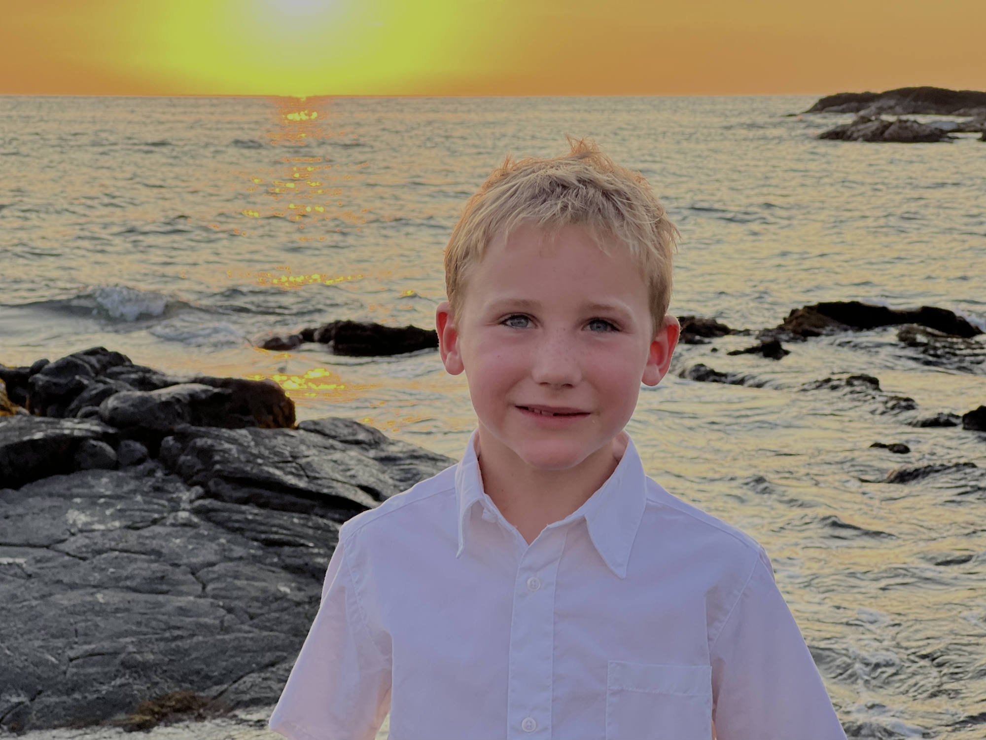 A young boy in a white button-down shirt smiles for a portrait on a rocky shoreline. Behind him, the sun sets over the ocean, casting a bright orange glow across the water and sky.