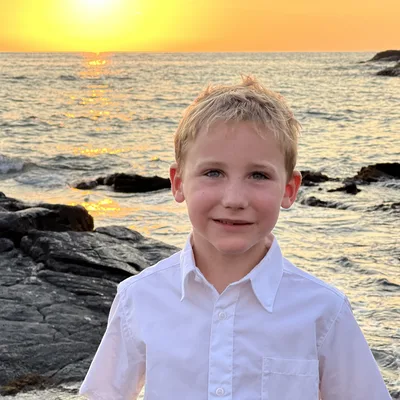 A young boy in a white button-down shirt smiles for a portrait on a rocky shoreline. Behind him, the sun sets over the ocean, casting a bright orange glow across the water and sky.