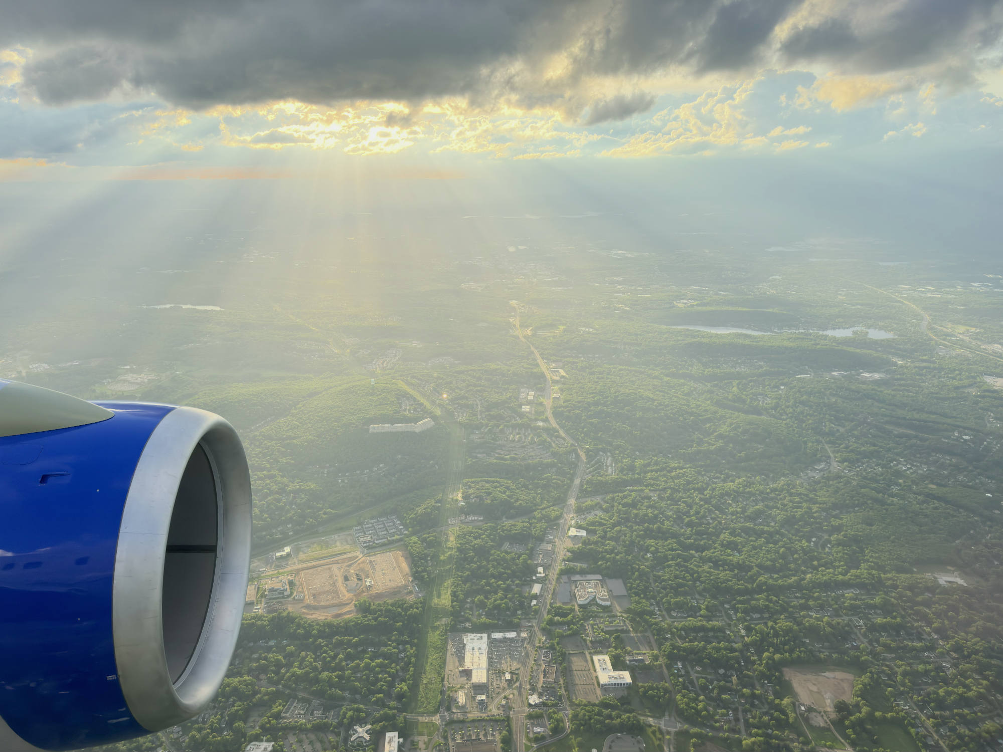An aerial view from an airplane window shows a blue engine in the foreground, looking out over a green landscape of forests and towns. Sunbeams break through dark storm clouds in the sky, illuminating the ground below.