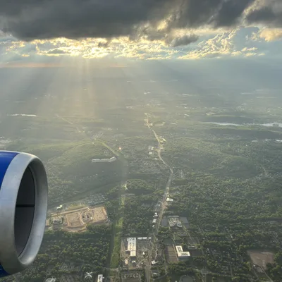 An aerial view from an airplane window shows a blue engine in the foreground, looking out over a green landscape of forests and towns. Sunbeams break through dark storm clouds in the sky, illuminating the ground below.