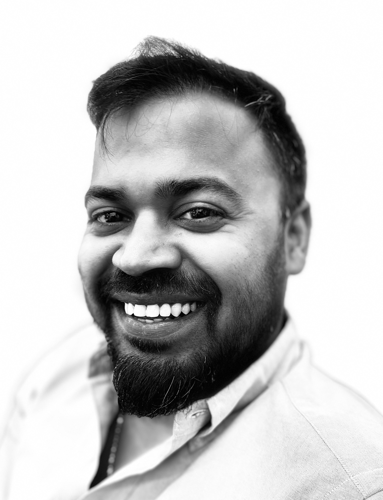 A black and white, close-up portrait of a smiling man with a beard and mustache. He is wearing a collared shirt and posing against a plain white background.