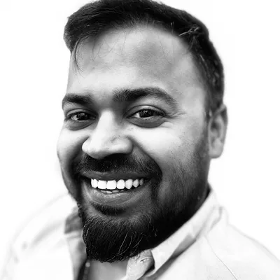 A black and white, close-up portrait of a smiling man with a beard and mustache. He is wearing a collared shirt and posing against a plain white background.