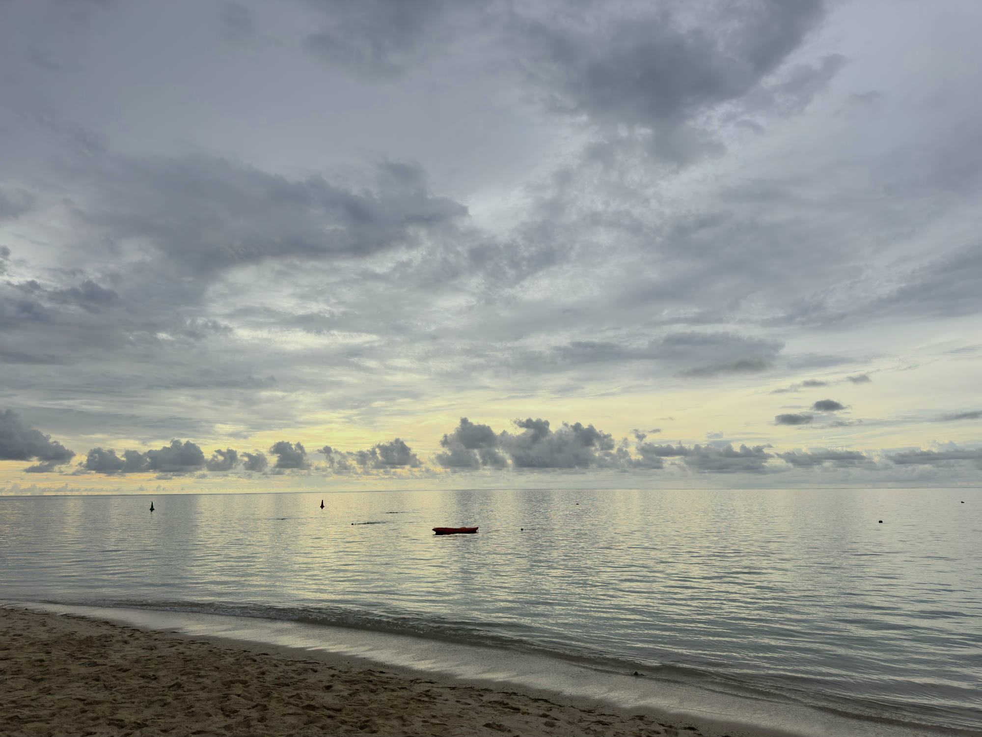 A wide shot of a sandy beach looking out over a calm ocean under a dramatic, cloudy sky at dusk. A small red kayak floats on the water near the shore, with a few distant buoys visible on the horizon.