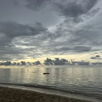 A wide shot of a sandy beach looking out over a calm ocean under a dramatic, cloudy sky at dusk. A small red kayak floats on the water near the shore, with a few distant buoys visible on the horizon.