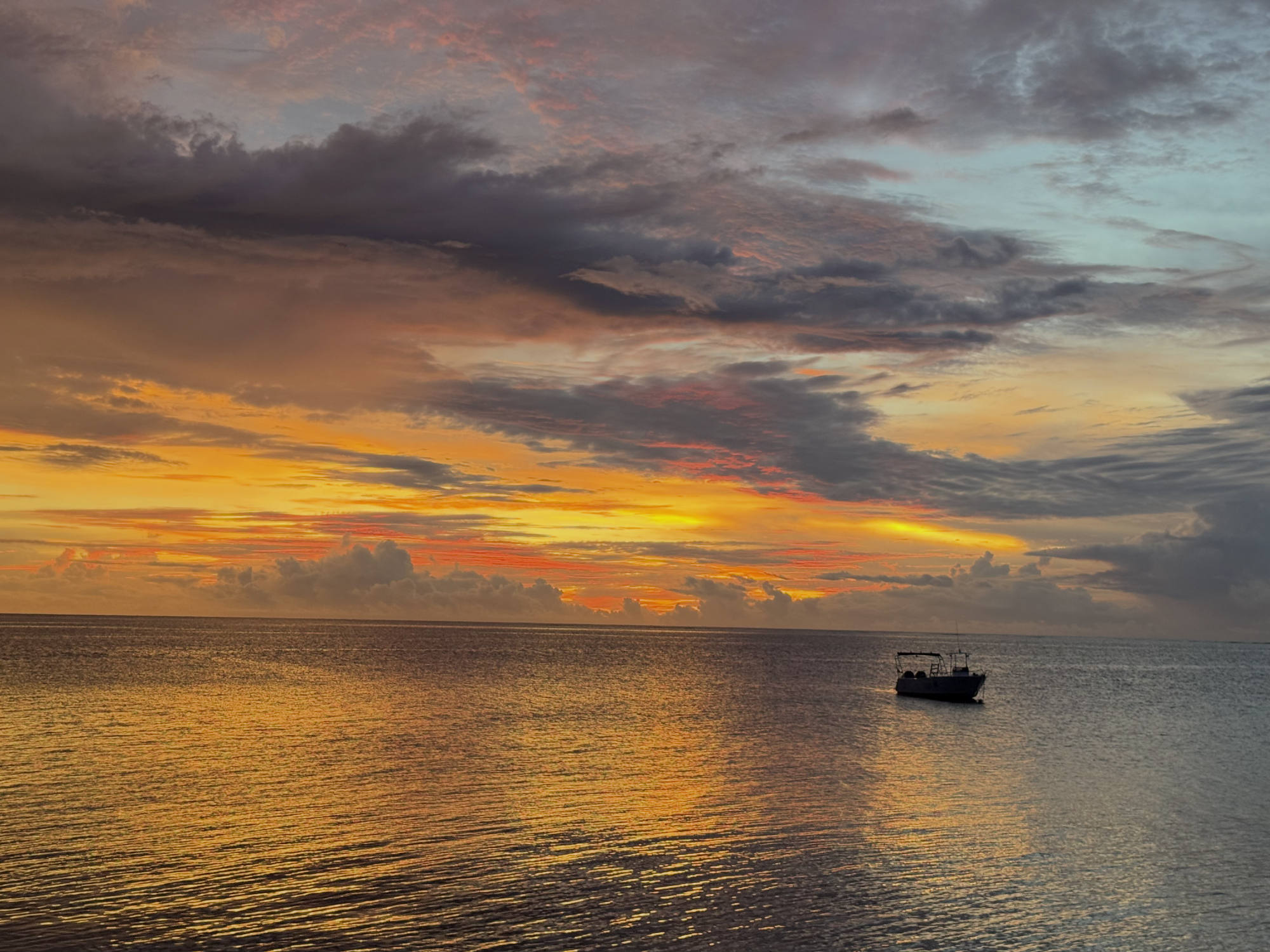 A small boat floats on calm ocean waters under a dramatic sunset sky filled with orange, yellow, and dark grey clouds. The vibrant colors of the sunset reflect across the surface of the water.