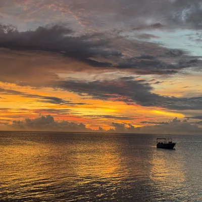A small boat floats on calm ocean waters under a dramatic sunset sky filled with orange, yellow, and dark grey clouds. The vibrant colors of the sunset reflect across the surface of the water.
