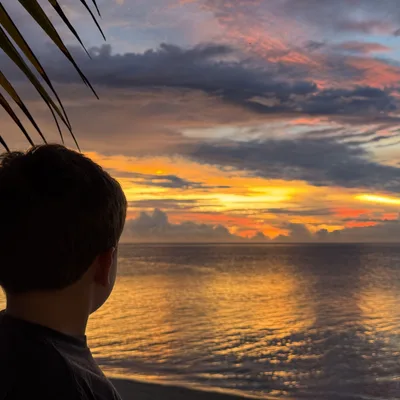A boy is seen from behind, looking out at a vibrant sunset over the ocean from a beach. The sky is filled with colorful orange, yellow, and pink clouds, which are reflected in the calm water below.