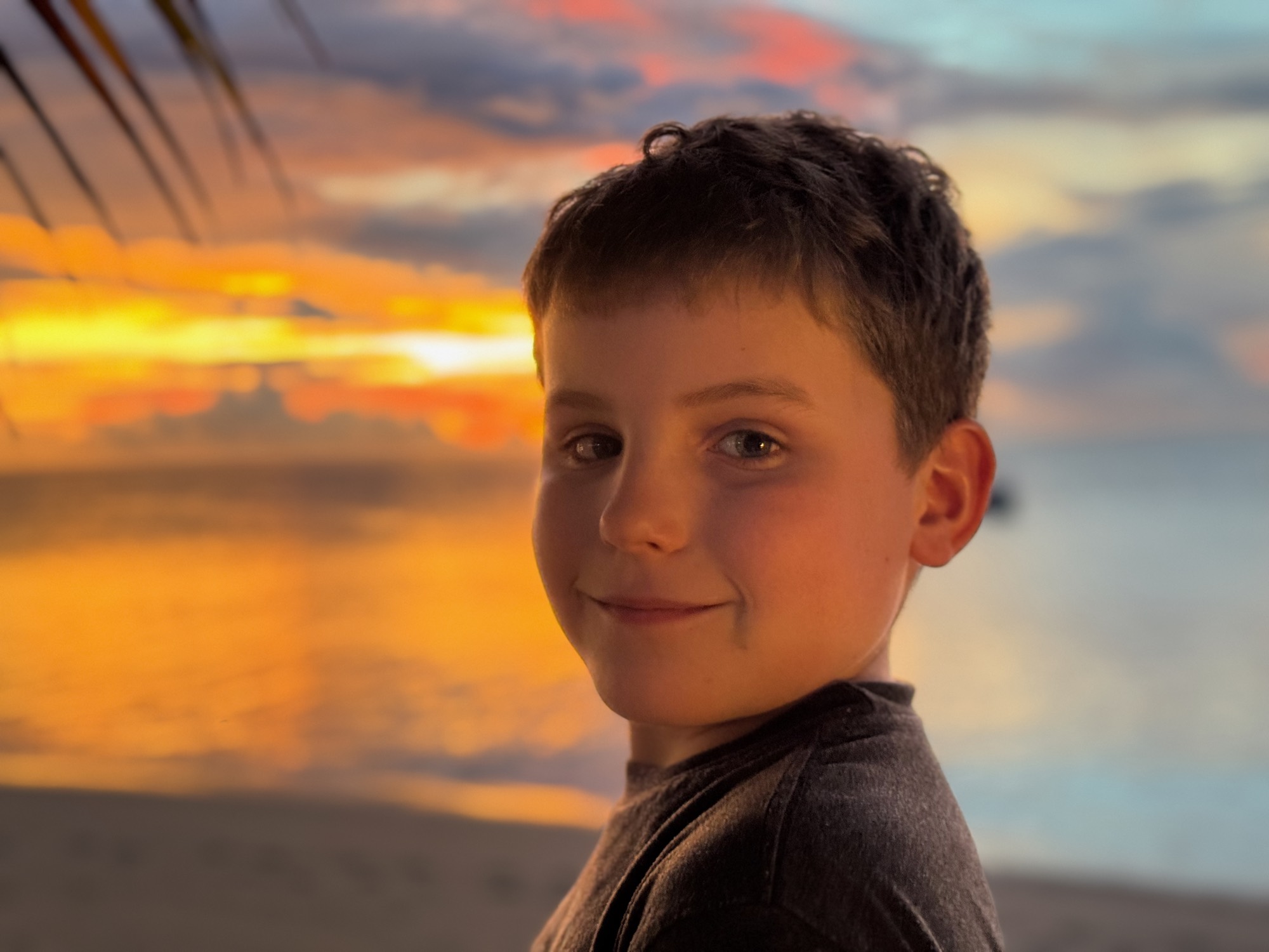 A close-up portrait of a smiling young boy looking back over his shoulder on a sandy beach. In the background, a vibrant orange and pink sunset reflects over the calm ocean water.