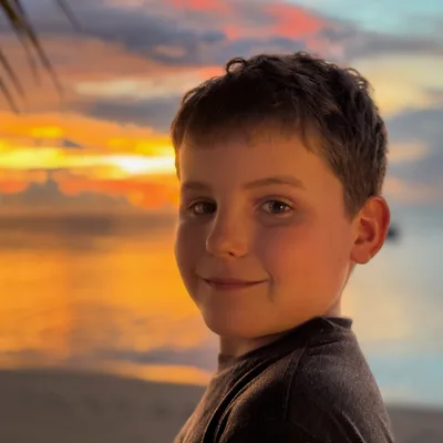 A close-up portrait of a smiling young boy looking back over his shoulder on a sandy beach. In the background, a vibrant orange and pink sunset reflects over the calm ocean water.