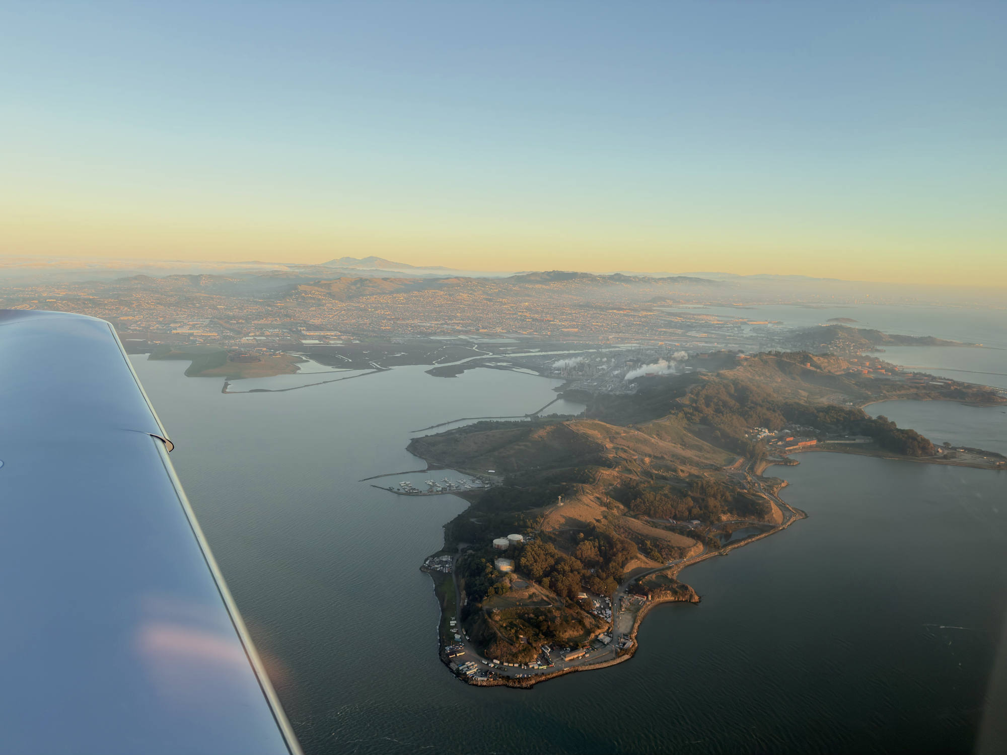 An aerial view from an airplane window captures a peninsula and surrounding bay at sunset, with a city and mountains visible in the background.