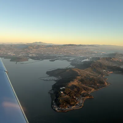 An aerial view from an airplane window captures a peninsula and surrounding bay at sunset, with a city and mountains visible in the background.