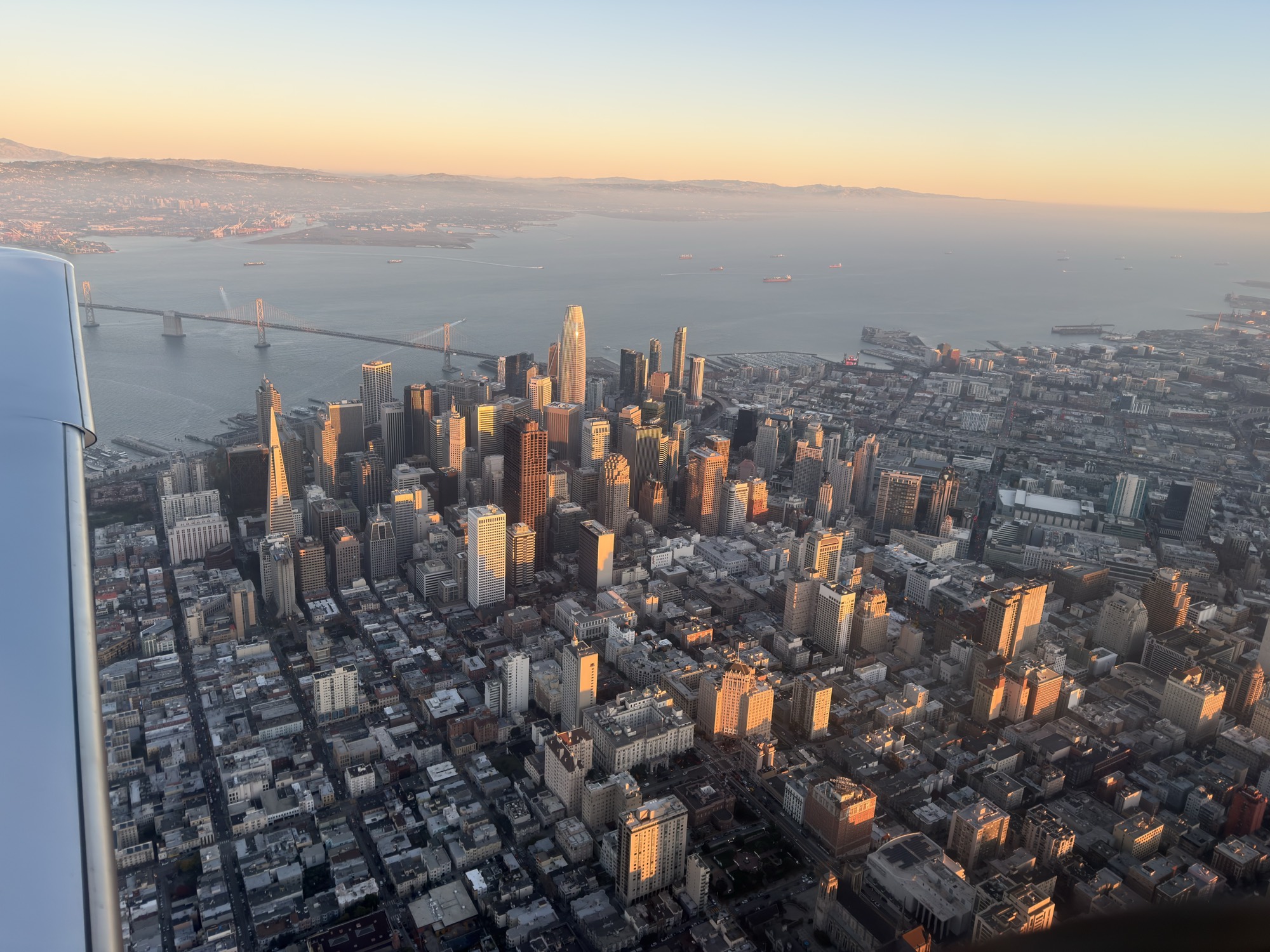 An aerial view from an airplane window captures the San Francisco skyline and the Bay Bridge illuminated by the warm glow of a sunset. The dense cityscape of skyscrapers and surrounding neighborhoods stretches toward the water, with distant hills visible under a clear, fading sky.