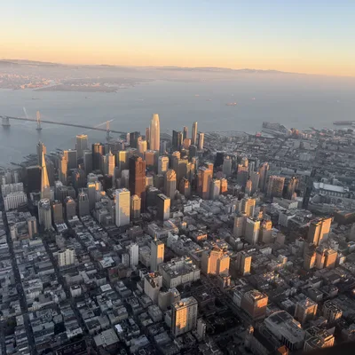 An aerial view from an airplane window captures the San Francisco skyline and the Bay Bridge illuminated by the warm glow of a sunset. The dense cityscape of skyscrapers and surrounding neighborhoods stretches toward the water, with distant hills visible under a clear, fading sky.