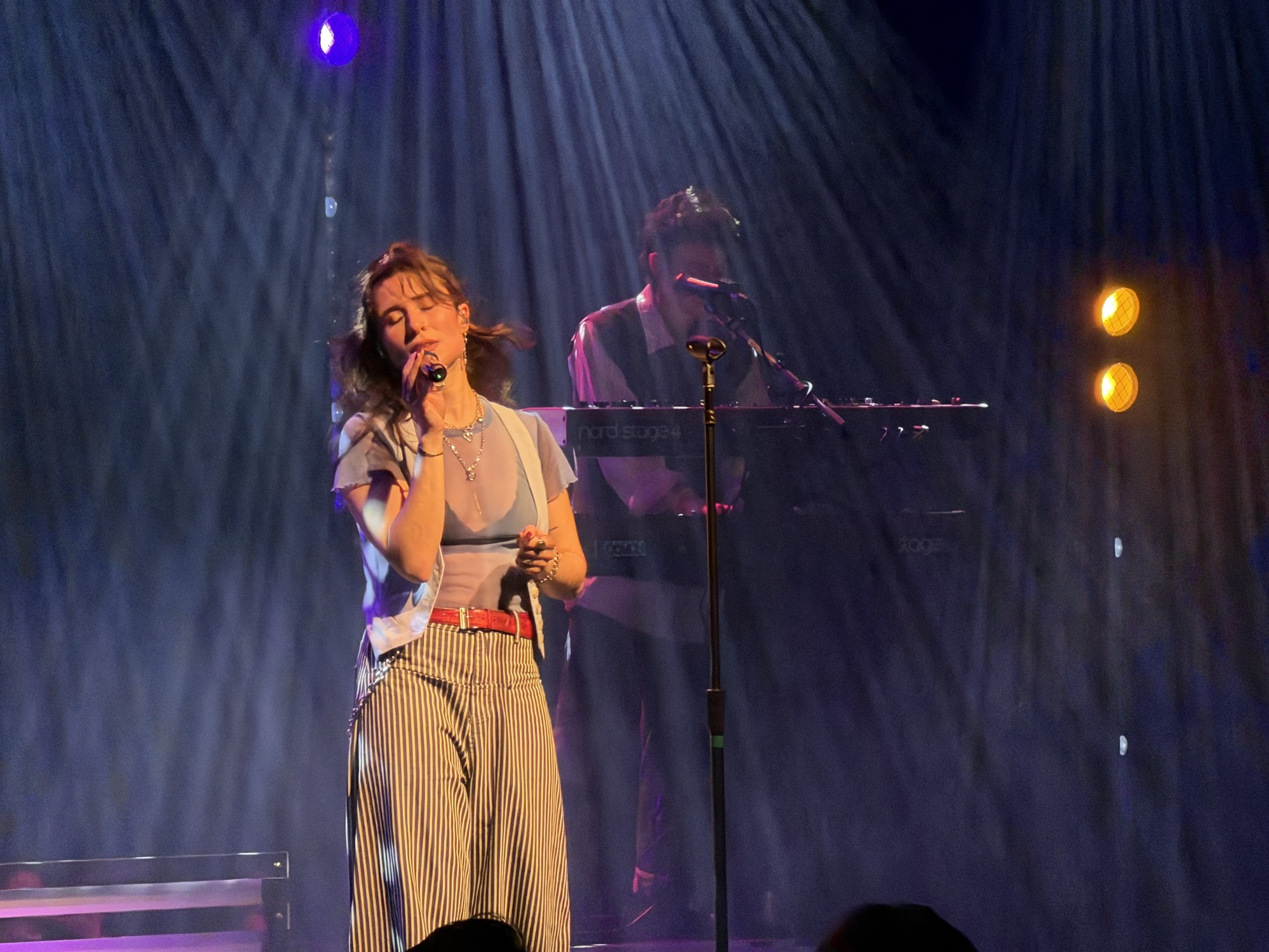 A female vocalist sings into a microphone on a dimly lit stage with blue and orange spotlights, while a keyboardist performs in the background.