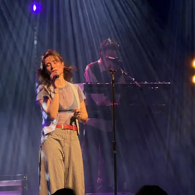 A female vocalist sings into a microphone on a dimly lit stage with blue and orange spotlights, while a keyboardist performs in the background.