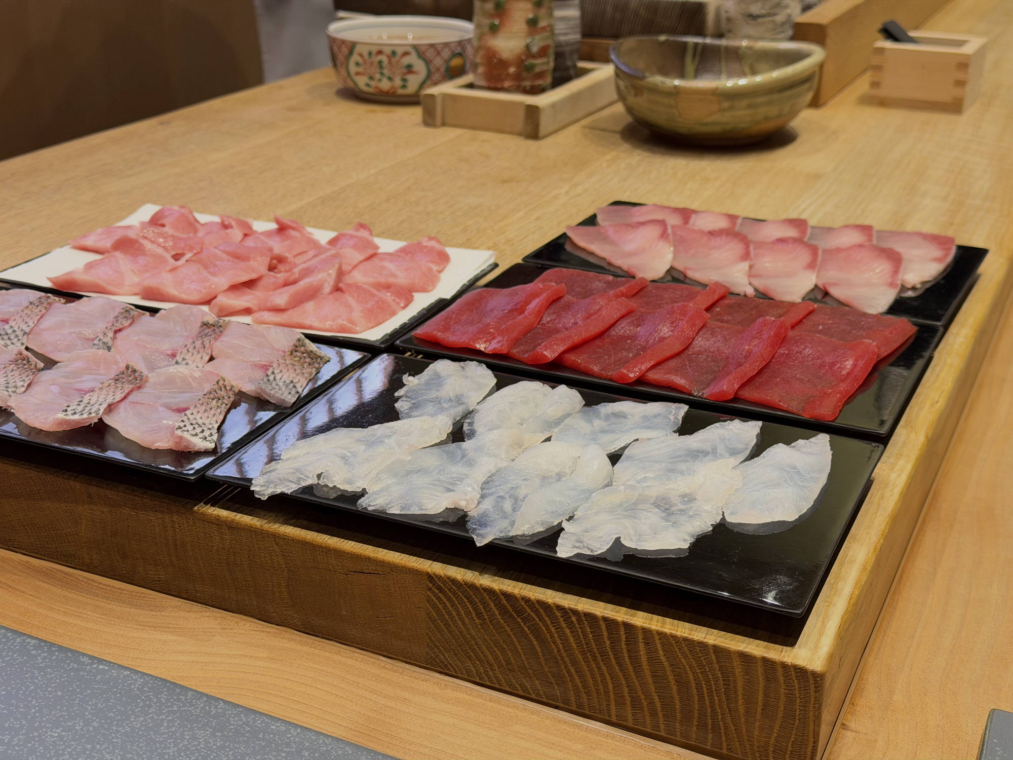 An assortment of sliced raw fish, prepared for sashimi or sushi, is arranged on black and white plates inside a wooden tray. The tray sits on a light-colored wooden counter in a restaurant setting, with small bowls visible in the background.