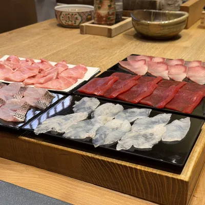 An assortment of sliced raw fish, prepared for sashimi or sushi, is arranged on black and white plates inside a wooden tray. The tray sits on a light-colored wooden counter in a restaurant setting, with small bowls visible in the background.