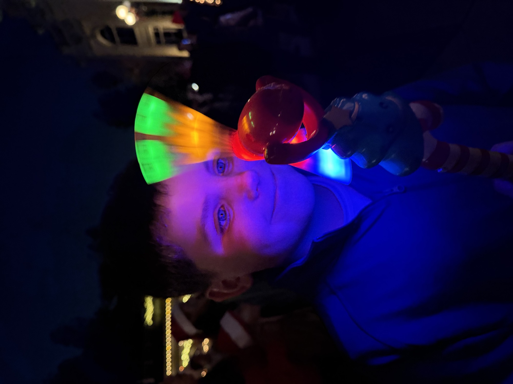 A young boy smiles as his face is illuminated by a spinning, light-up toy wand he is holding. The scene takes place outdoors at night, with blurred holiday lights and a crowd visible in the background.